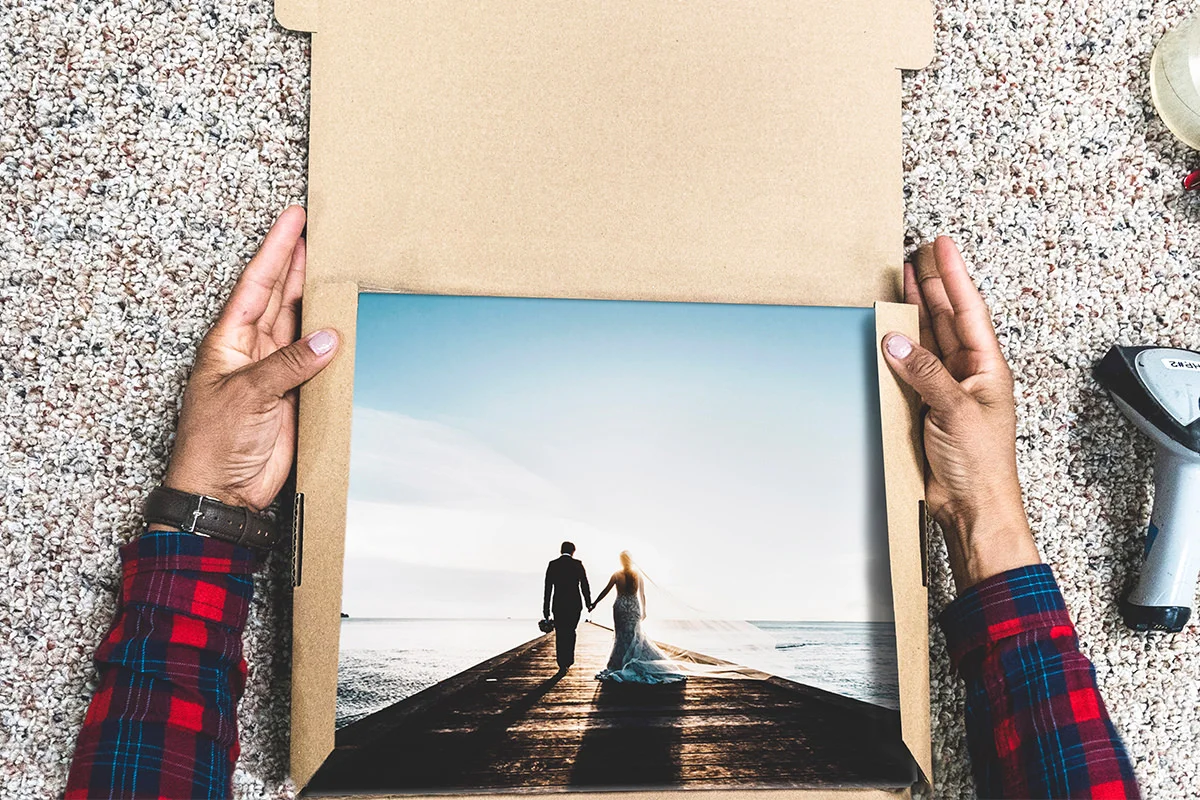 Hands holding open a cardboard box revealing a photo of a couple in formal wear walking on a pier at sunset.