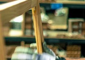 Close-up of a wooden dowel or leg in a workshop, with blurred shelves in the background.